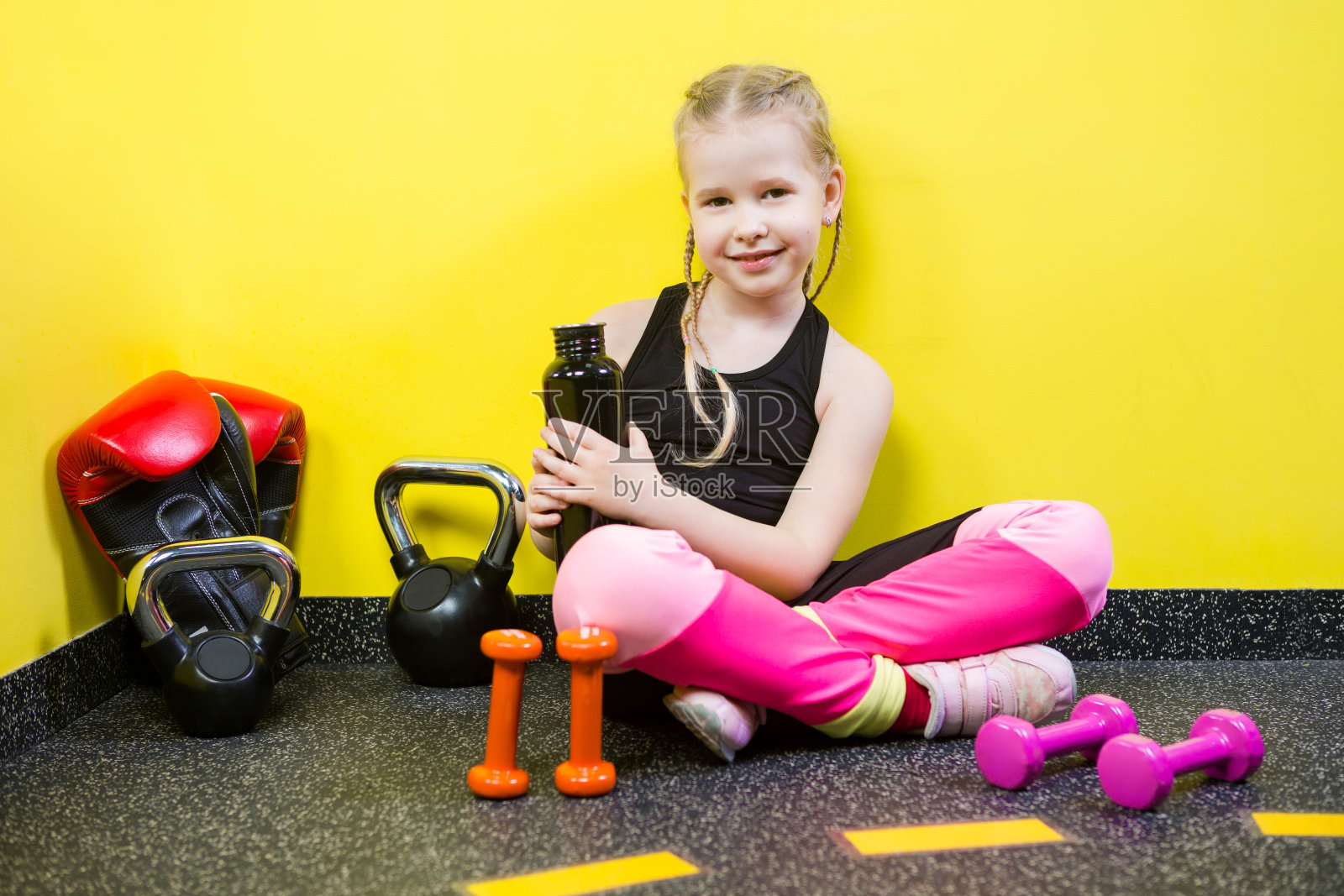 主题运动与健康儿童。小白种女孩坐在休息休息的地板健身房拿着水壶，喝着水干渴。运动员哑铃设备体操健身背景黄墙照片摄影图片