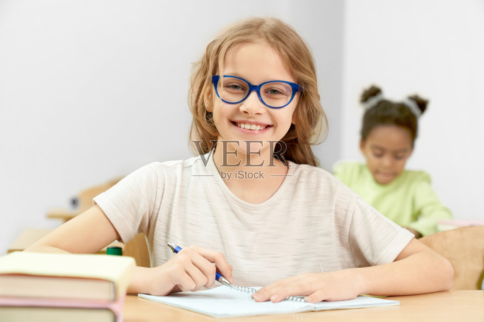 开朗聪明的女孩看着相机和摆姿势，在课堂上做任务。女学生坐在书桌前，写笔记本和学习。学习过程的概念。照片摄影图片