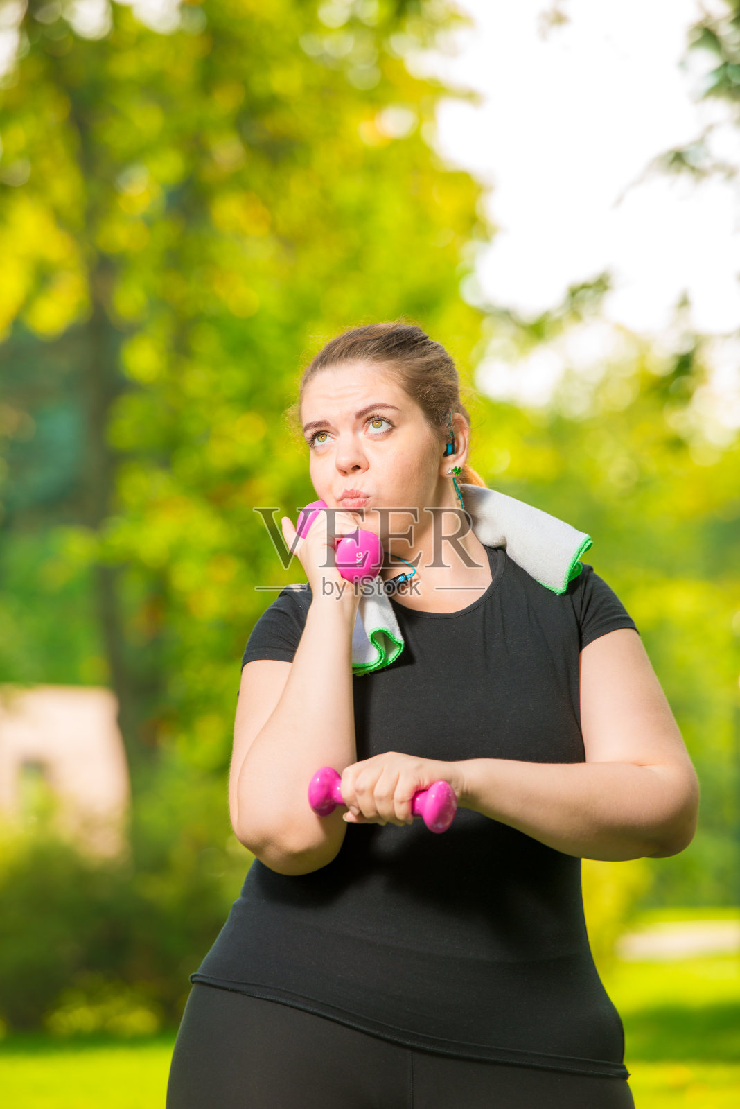 细心的大码模特戴着耳机听音乐，在夏天的公园里练习哑铃照片摄影图片