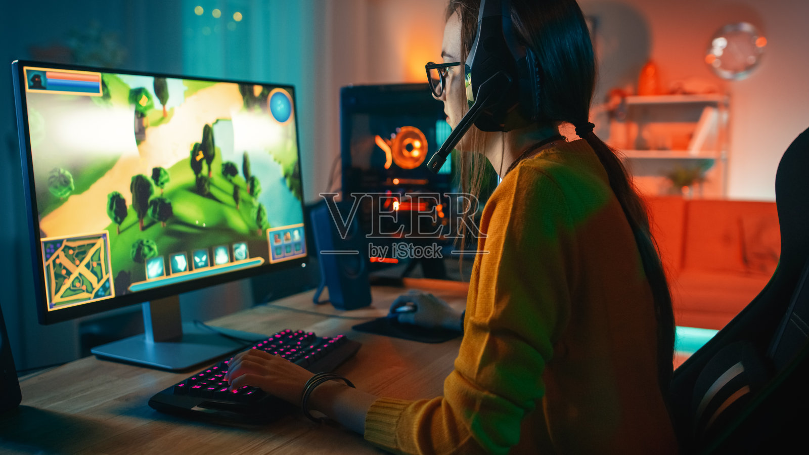 戴着眼镜戴着麦克风在个人电脑上玩在线策略视频游戏的兴奋和专注的玩家女孩。房间和电脑有彩色暖霓虹灯Led灯。温馨的家庭之夜。照片摄影图片