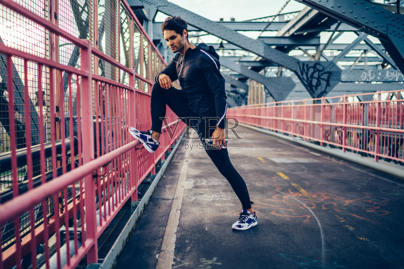 年轻人在活动中戴着耳机听音乐，在现代桥训练前热身。穿着黑色运动服和运动运动鞋的运动男子在户外锻炼，同时享受音频歌曲照片摄影图片