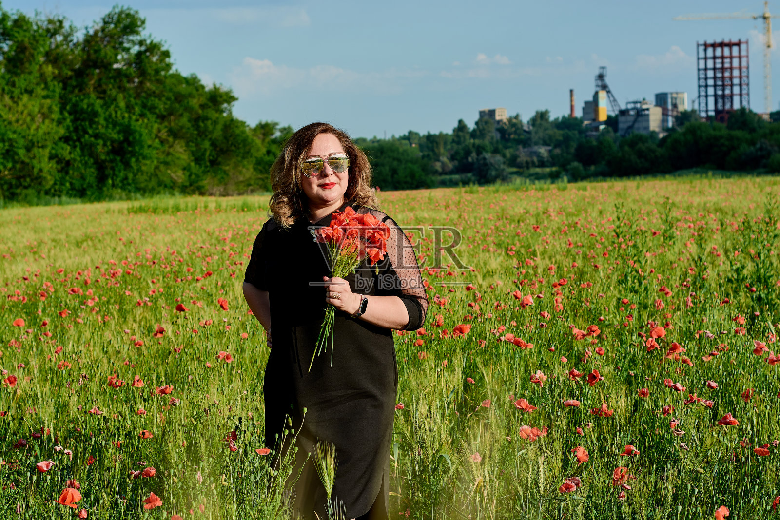 一个穿着黑色裙子的胖女人站在绿色的小麦和野罂粟地里。超重的胖女人。照片摄影图片