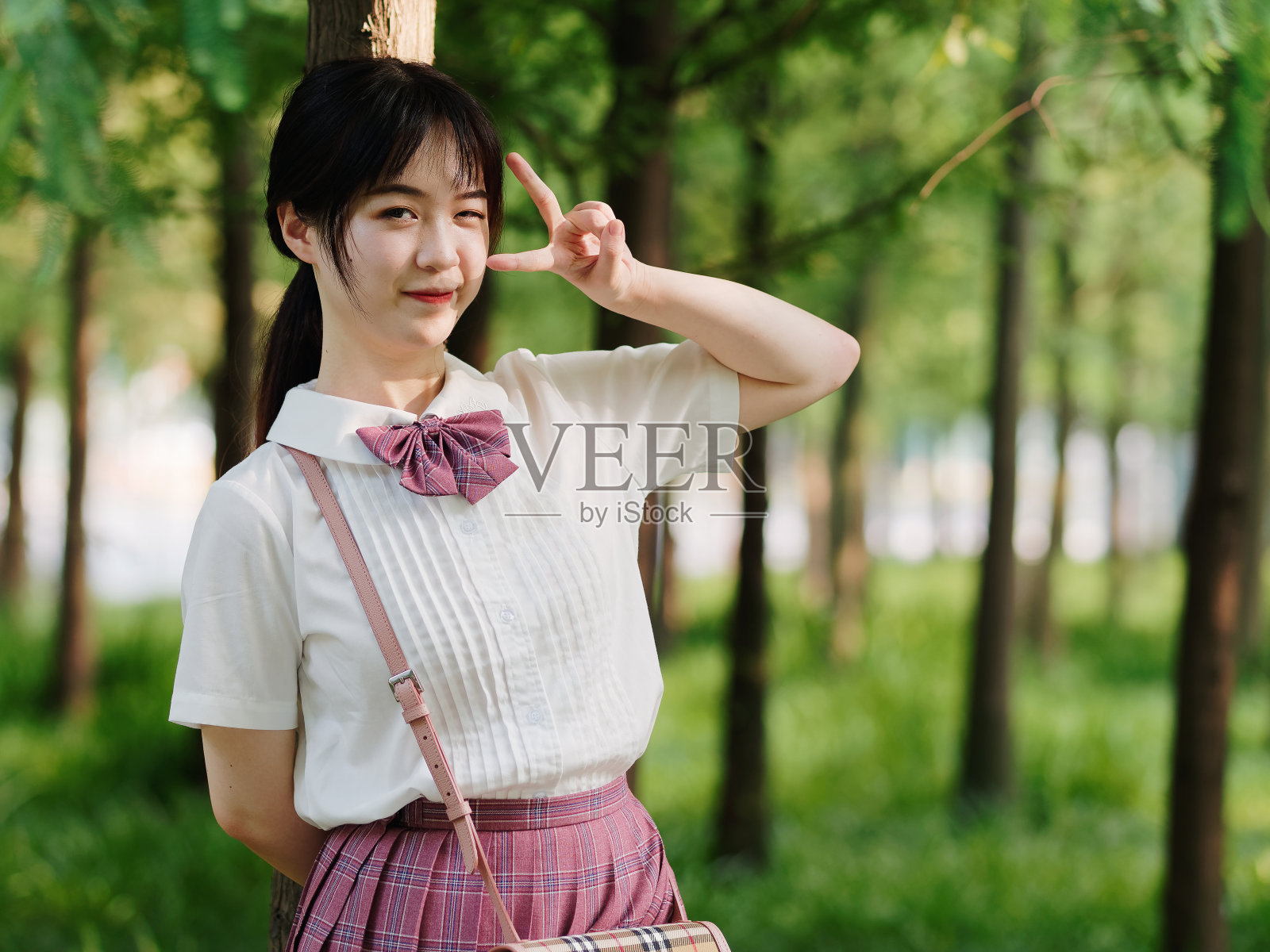 在阳光明媚的夏日森林里，美丽的中国女孩穿着白色衬衫和粉色裙子微笑，用剪刀手势做鬼脸，美丽，生活方式和夏天的概念。照片摄影图片