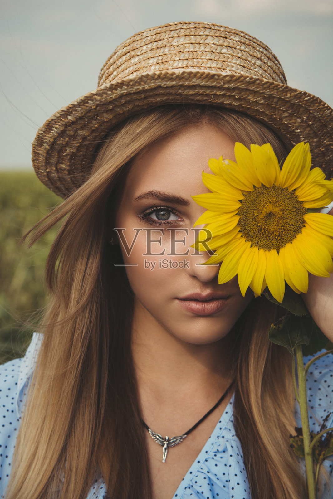 一头金发的年轻女子戴着草帽，穿着蓝色的衣服，站在向日葵地里。照片摄影图片