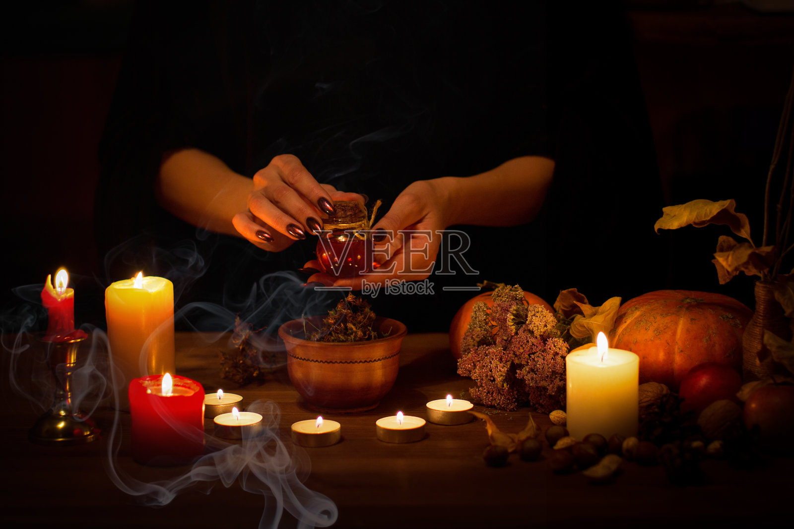 女巫制作了一些药剂，咒语和收集材料在祭坛上在黑暗中。女性的手用锋利的黑色指甲夹在蜡烛、南瓜、坚果、魔法药草、干叶子之间，选的重点，低调照片摄影图片