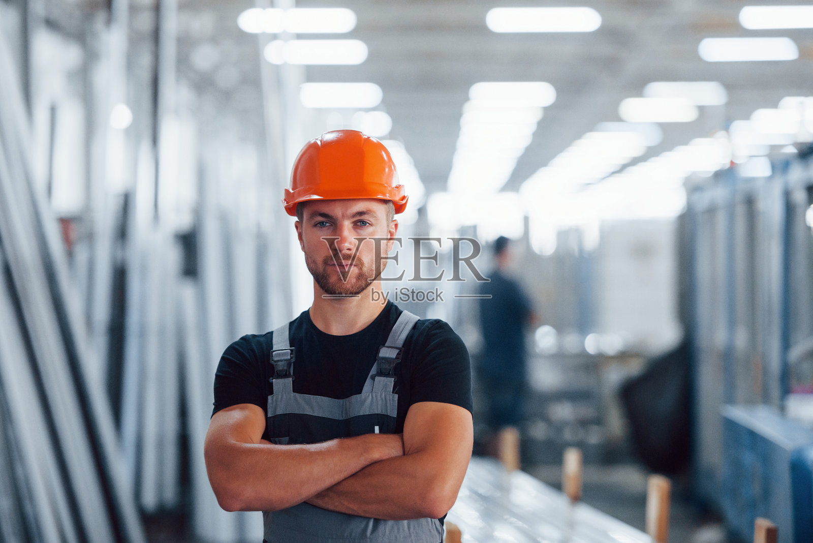 双臂交叉站着。男性工业工人在工厂室内的肖像。戴着橙色安全帽的年轻技术员照片摄影图片