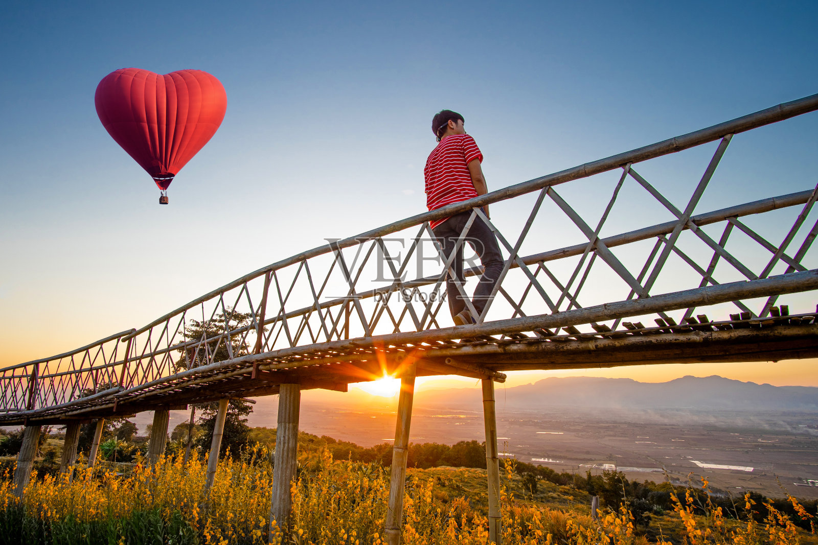 一名亚洲男子的剪影站在竹桥上，手捧心形红色热气球，夕阳西下。照片摄影图片