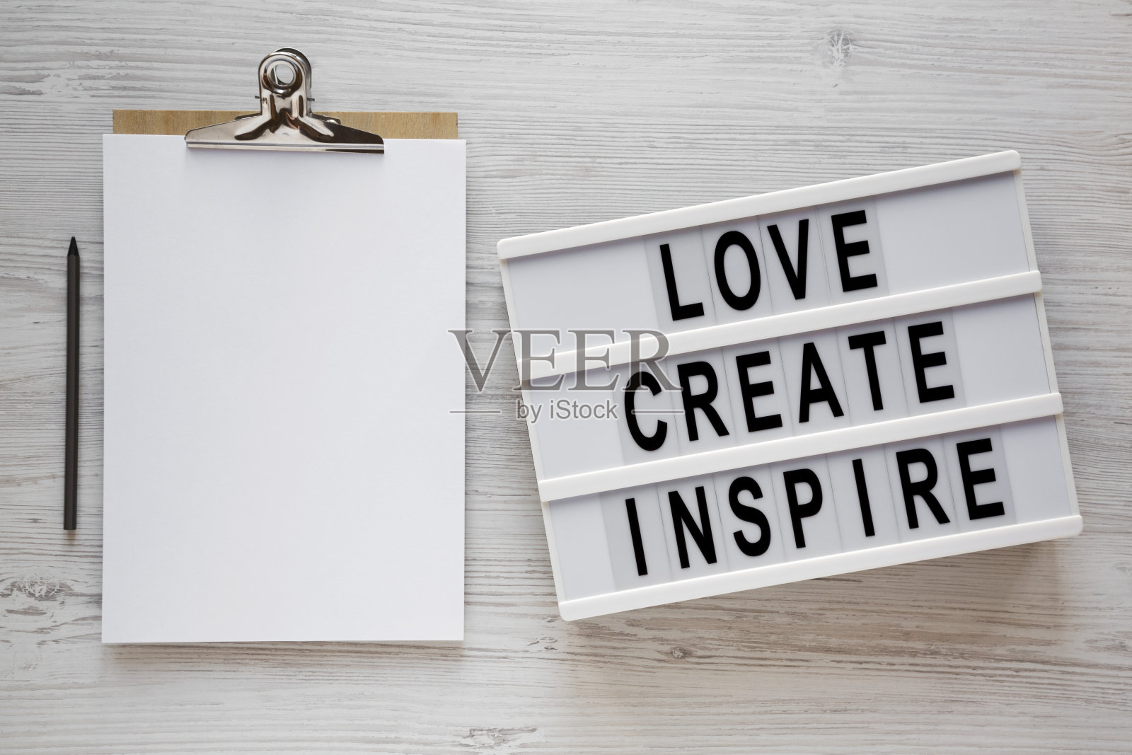 “爱创造灵感”的字样写在灯箱上，白色木质表面上有一张白纸的剪贴板，俯视图。头顶上，从上面看，是平躺着的。照片摄影图片