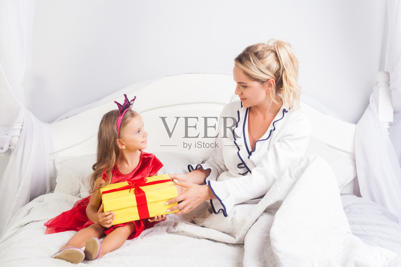 年轻的女人坐在床上，把礼物送给穿着鲜艳衣服、头戴皇冠的可爱小女孩照片摄影图片