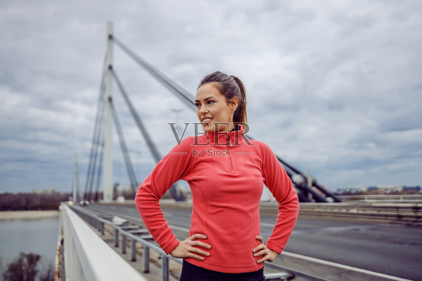 骄傲英俊的年轻运动黑发女子站在桥上，手放在臀部，在跑步中休息。城市生活。户外健身对多云天气的概念。照片摄影图片