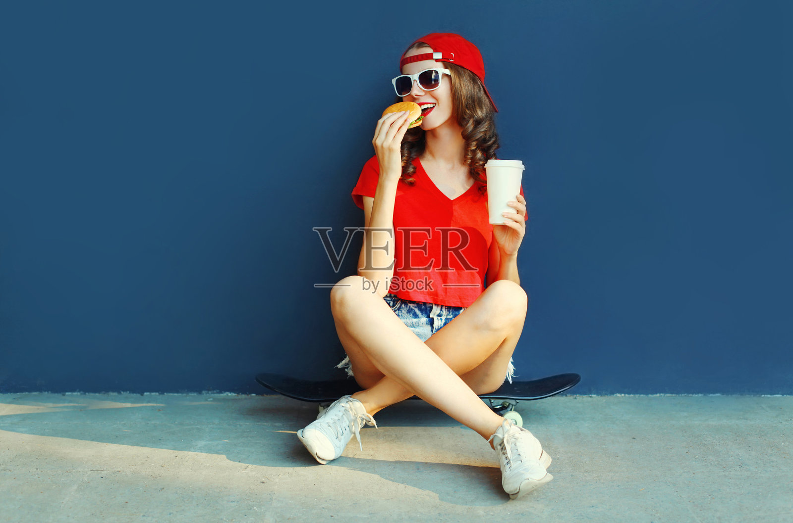酷酷的年轻女子与汉堡和咖啡杯坐在滑板戴着棒球帽，太阳镜在蓝色墙的背景照片摄影图片