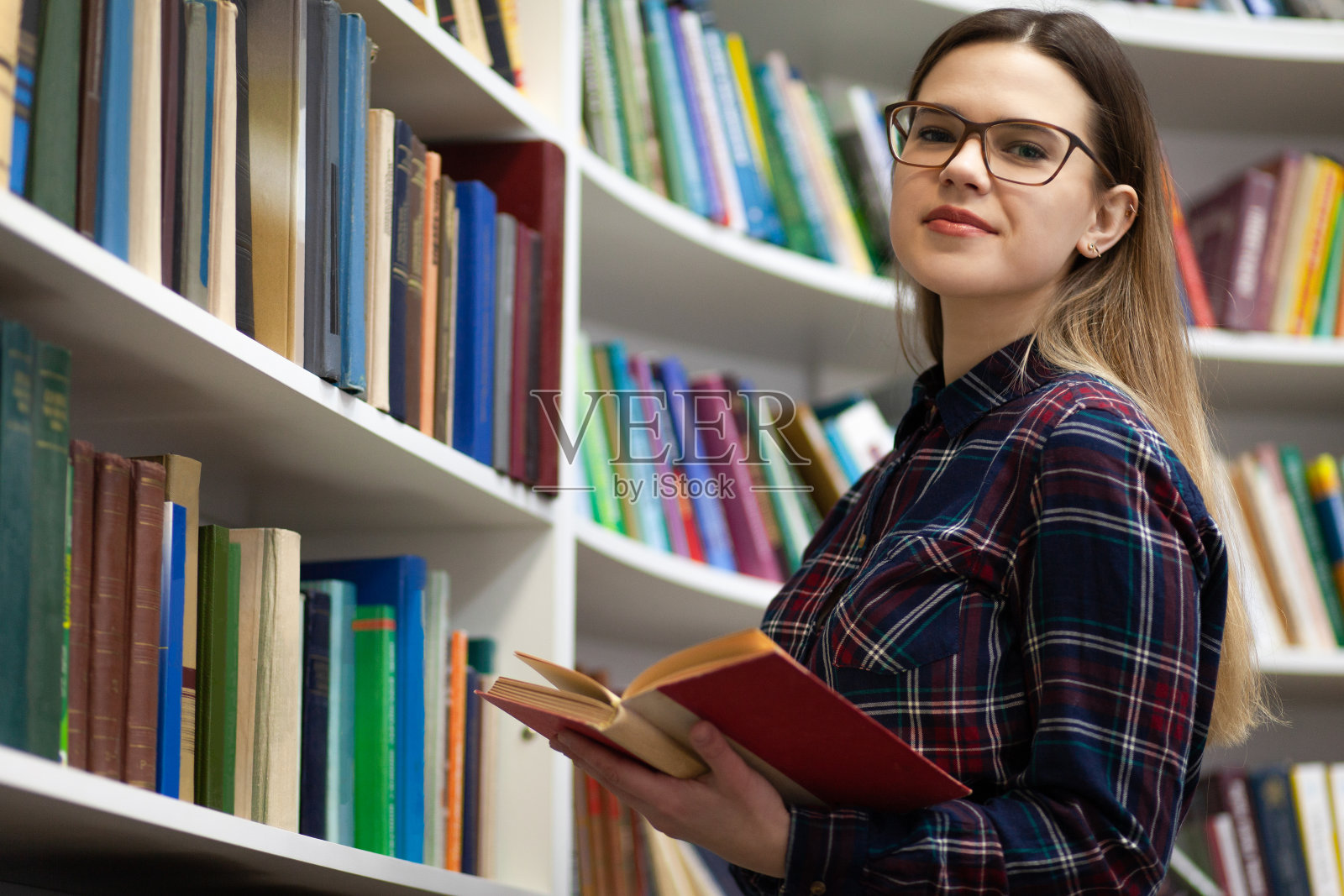 青少年手里拿着一本书站在图书馆里。一个戴眼镜的年轻学生在读一本书的肖像照片摄影图片
