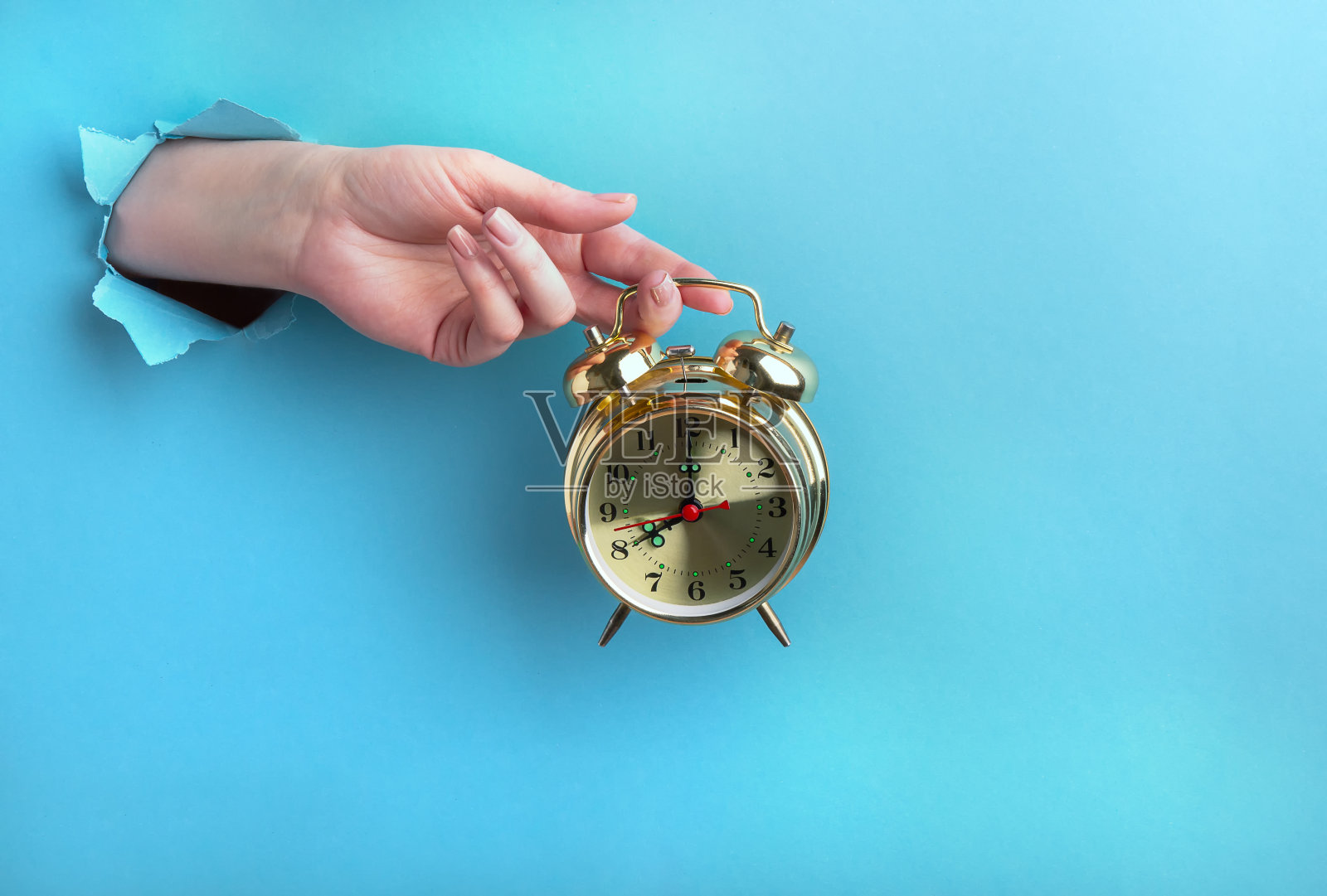 女性的手通过蓝色背景上的一个洞拿着一个金色的闹钟。,时间的概念照片摄影图片