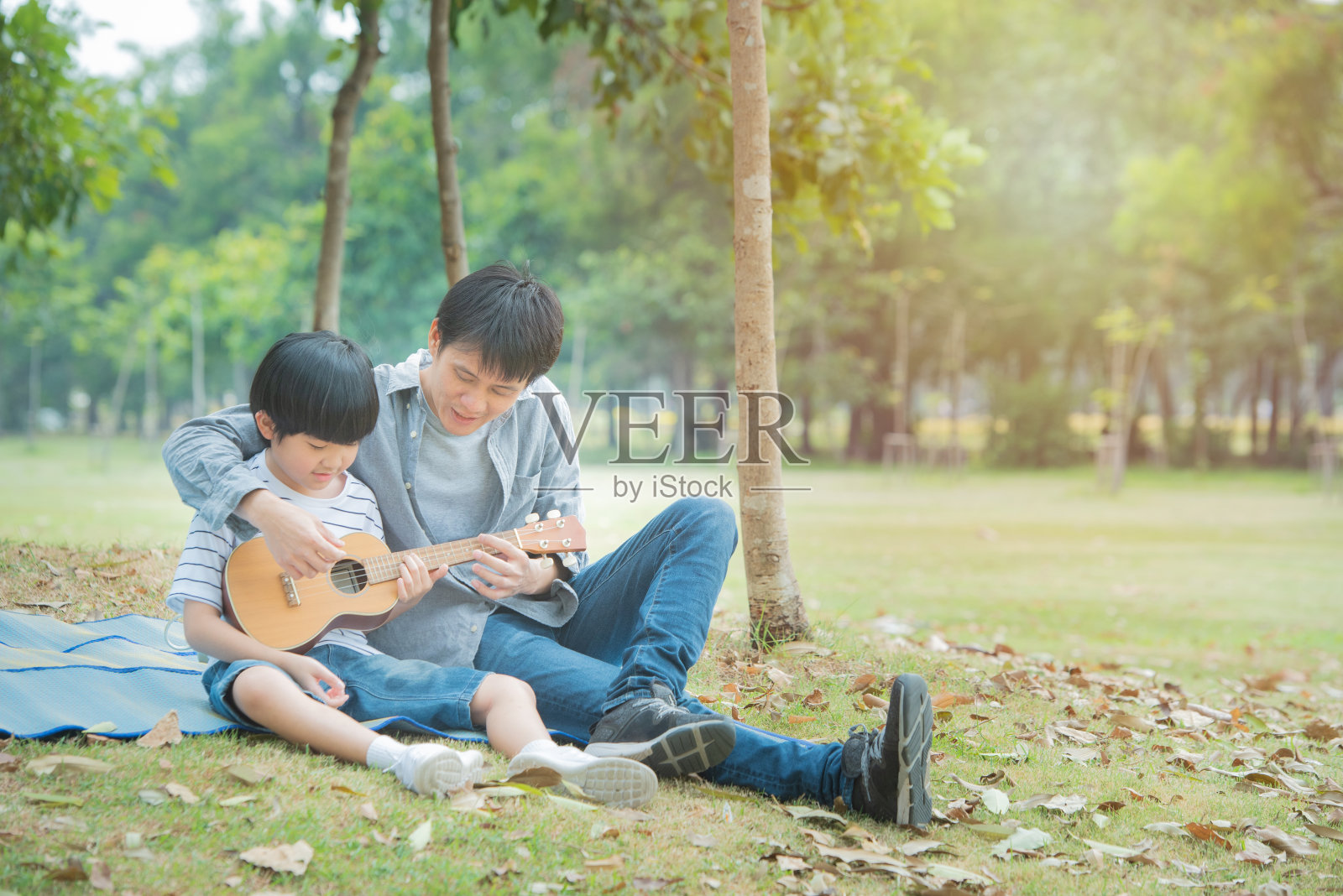 亚洲父亲教儿子在公共公园弹吉他，快乐的父母在户外花园野餐活动。照片摄影图片