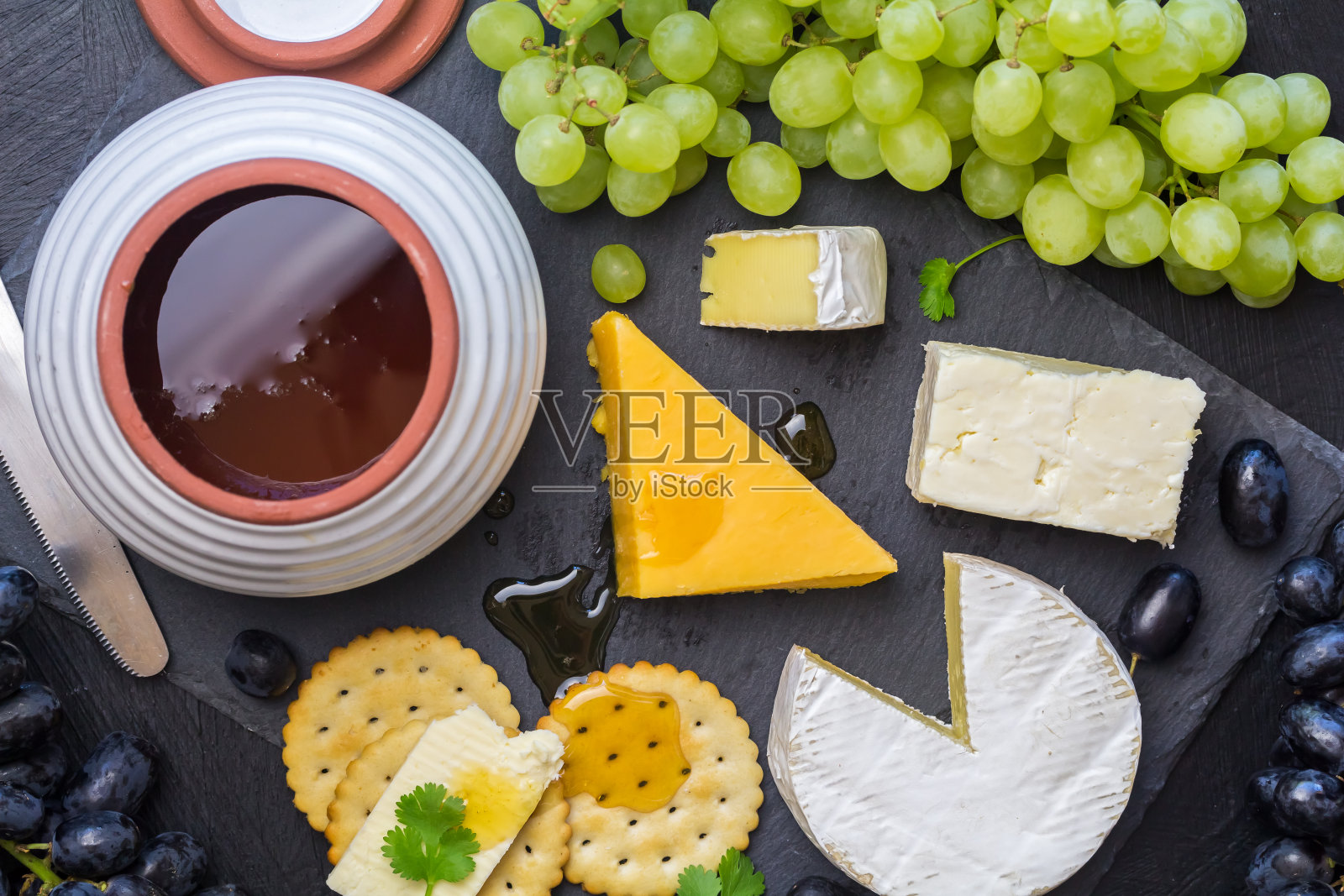奶酪板与各种奶酪，饼干，葡萄和蜂蜜罐在黑色石板的背景-上方的图像照片摄影图片