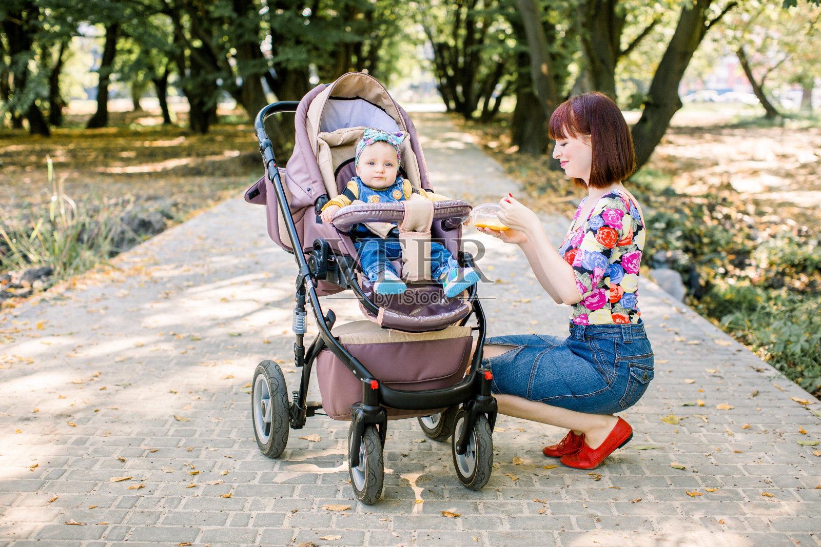 妈妈用勺子喂在公园里推着婴儿车走路的孩子。照片摄影图片
