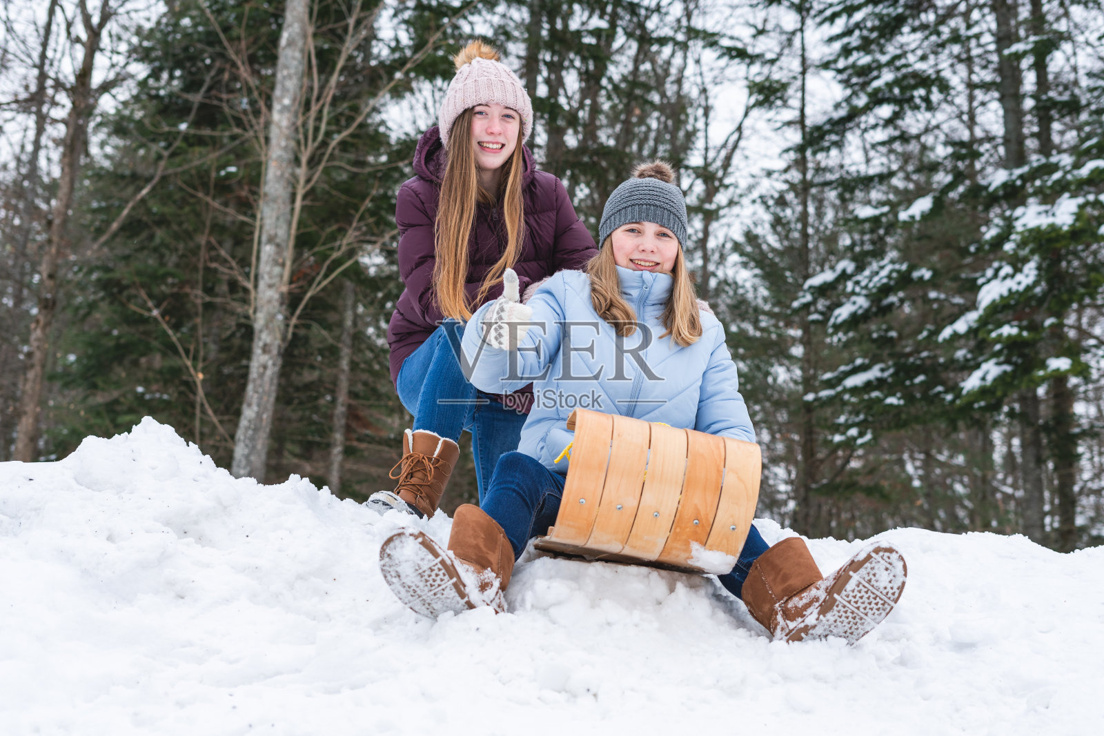 两个快乐的少女/朋友坐在一个木制雪橇/雪橇上，而在一个公园的雪覆盖的山的边缘和一个女孩举起她的拇指。照片摄影图片