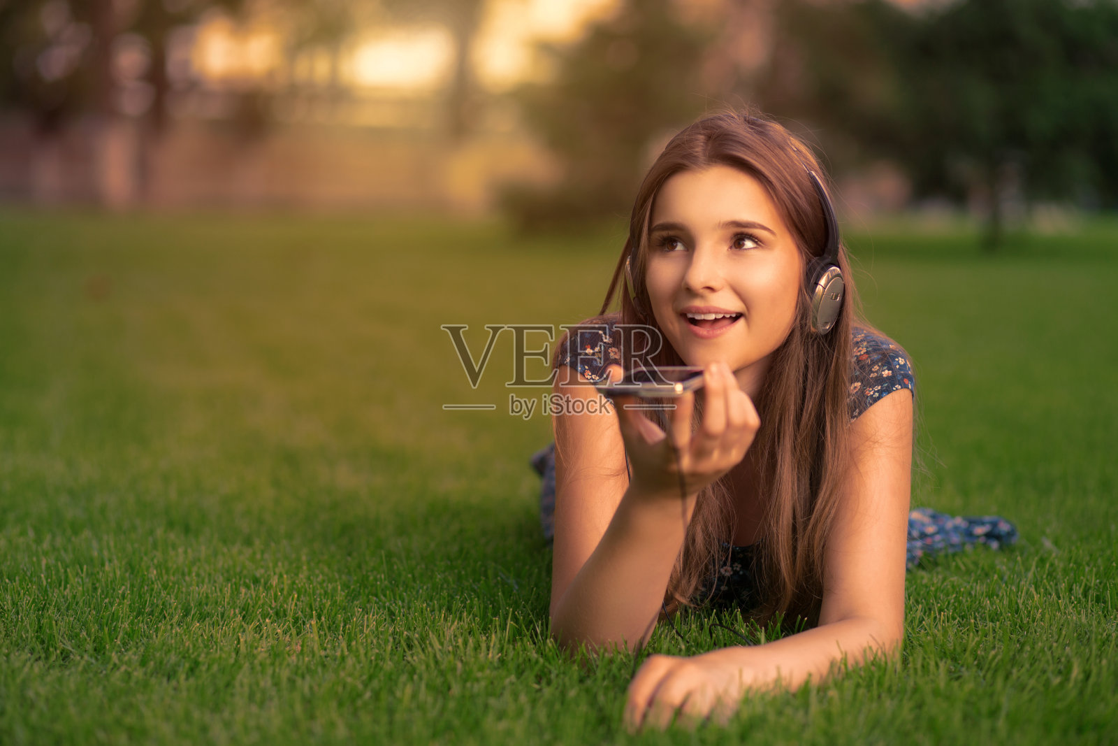 一名学生戴着耳机坐在公园的草地上，用智能手机进行语音识别记录笔记照片摄影图片
