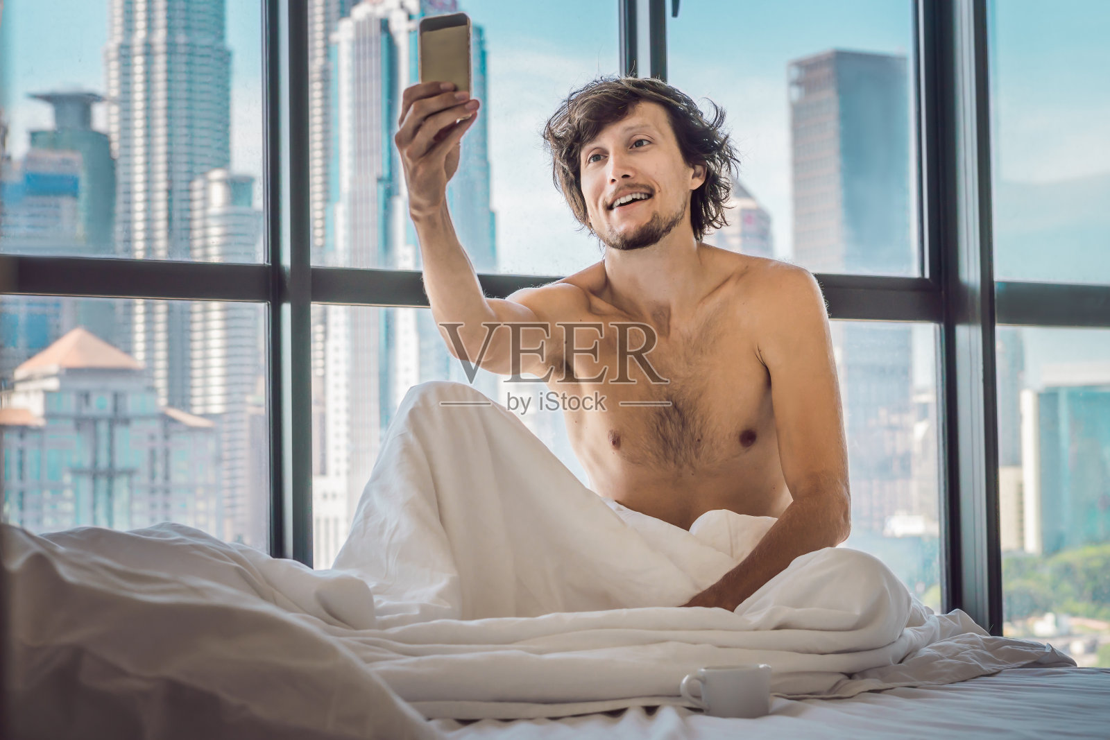一名男子早上在市中心的一间能看到摩天大楼的公寓里醒来，并使用智能手机。生活在喧嚣的大城市概念。没有足够的睡眠照片摄影图片