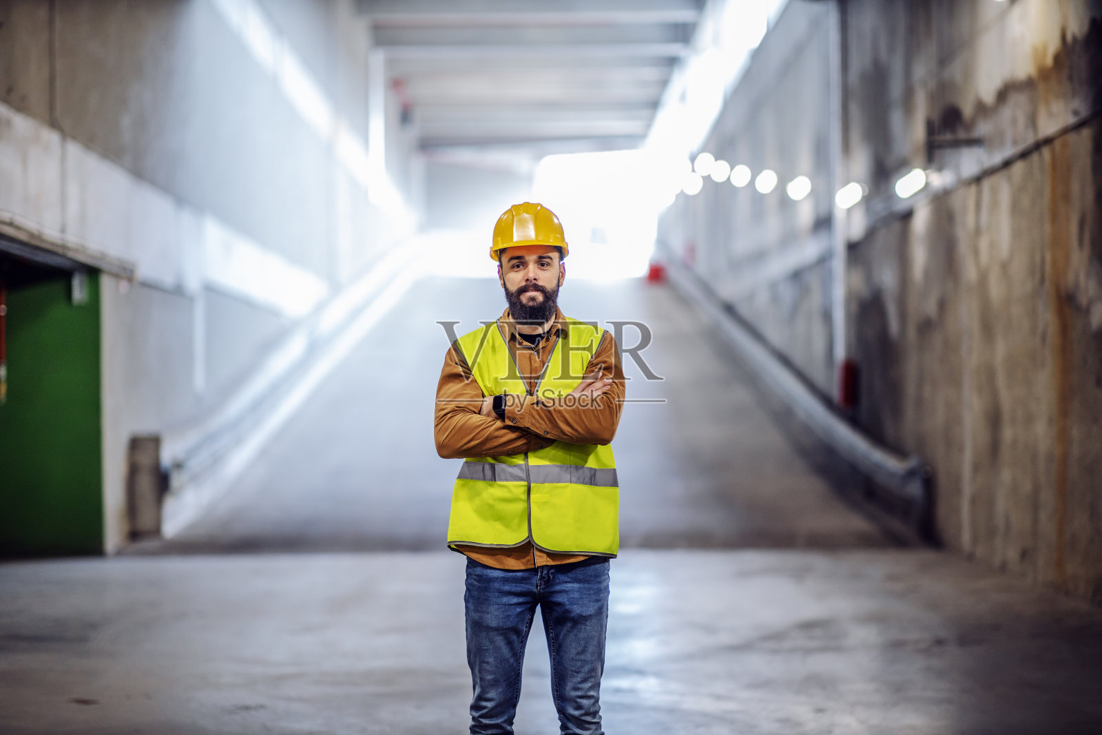 年轻有魅力的骄傲成功的胡子主管站在地下停车场的入口，在建设过程中双臂交叉。照片摄影图片