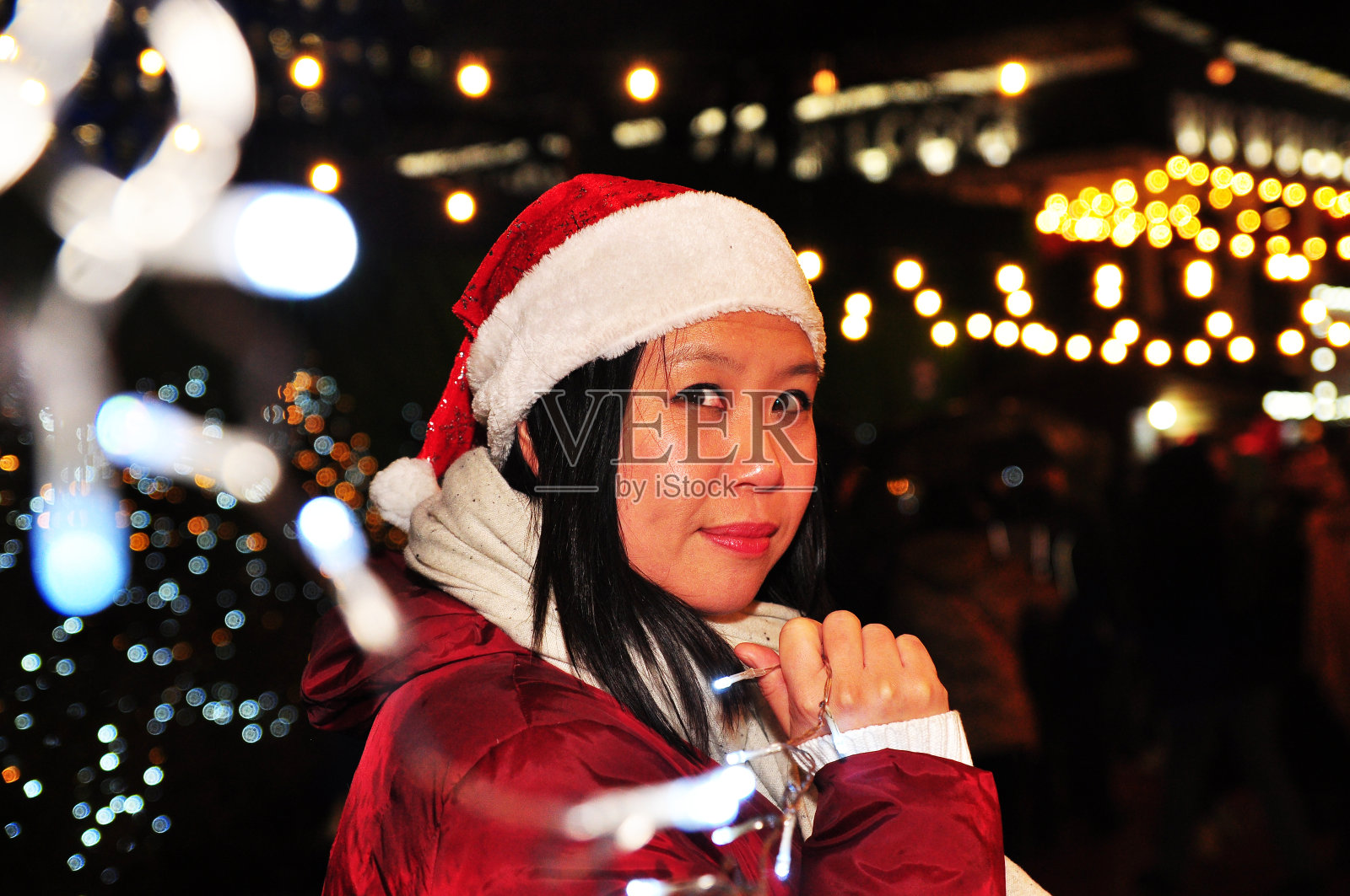 漂亮的亚洲女人拿着一串童话灯，戴着红色的圣诞帽和围巾，背景是明亮的圣诞灯在一个节日市场照片摄影图片