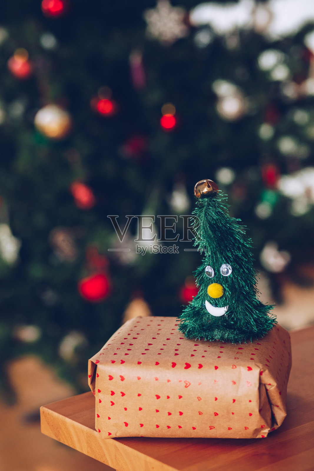 有趣的微笑的小圣诞玩具顶部包装的圣诞礼物在圣诞树前，在一个公寓室内的木桌上放置。照片摄影图片