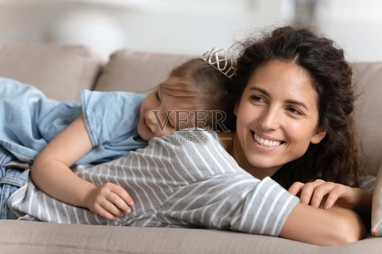 年轻的母亲微笑着躺在沙发上，女儿躺在她的背上。照片摄影图片