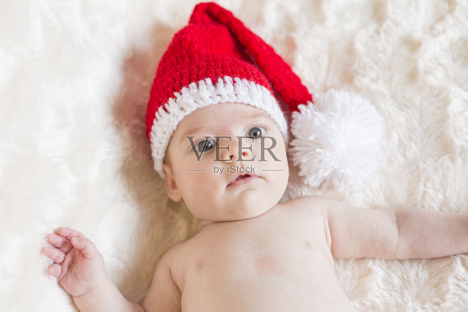 一个可爱的小女孩的肖像，醒着，看着相机。她戴着圣诞老人的帽子。白色的毯子背景照片摄影图片