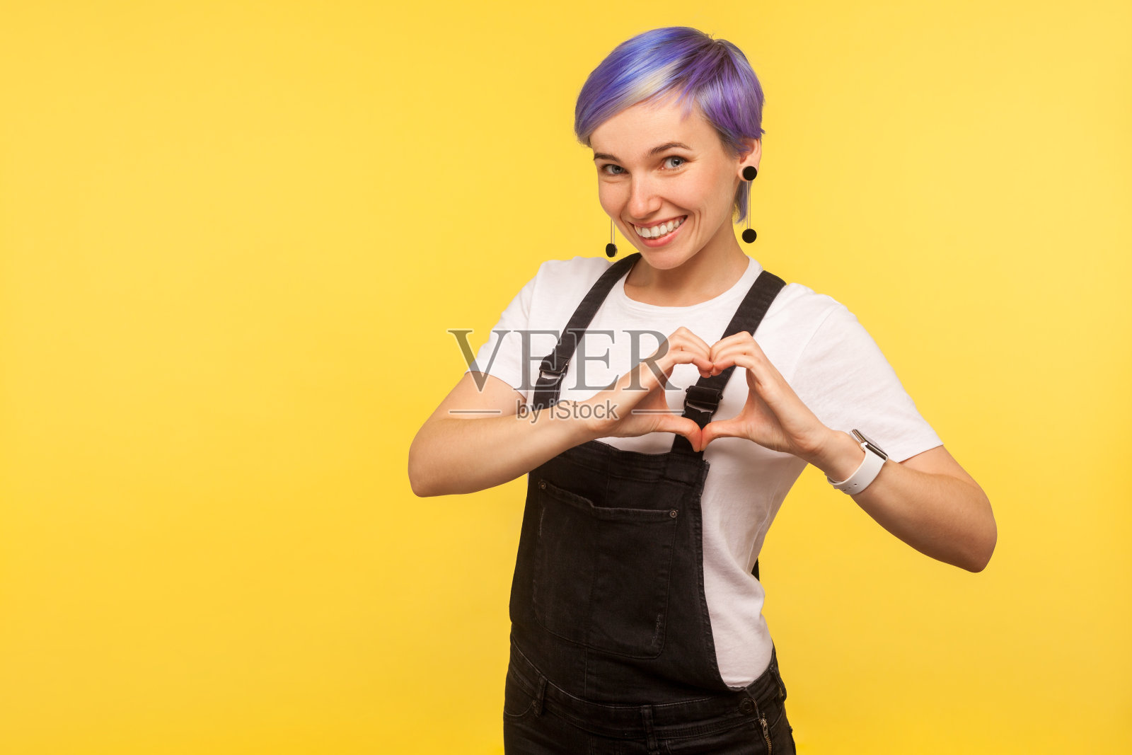 可爱的浪漫的嬉皮士女孩的肖像友好地微笑和显示心形符号与手。黄色背景，工作室拍摄照片摄影图片