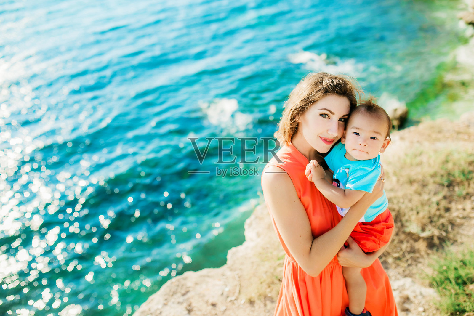 夏天的母亲在岩石和海岸的背景下拥抱着她的小儿子照片摄影图片