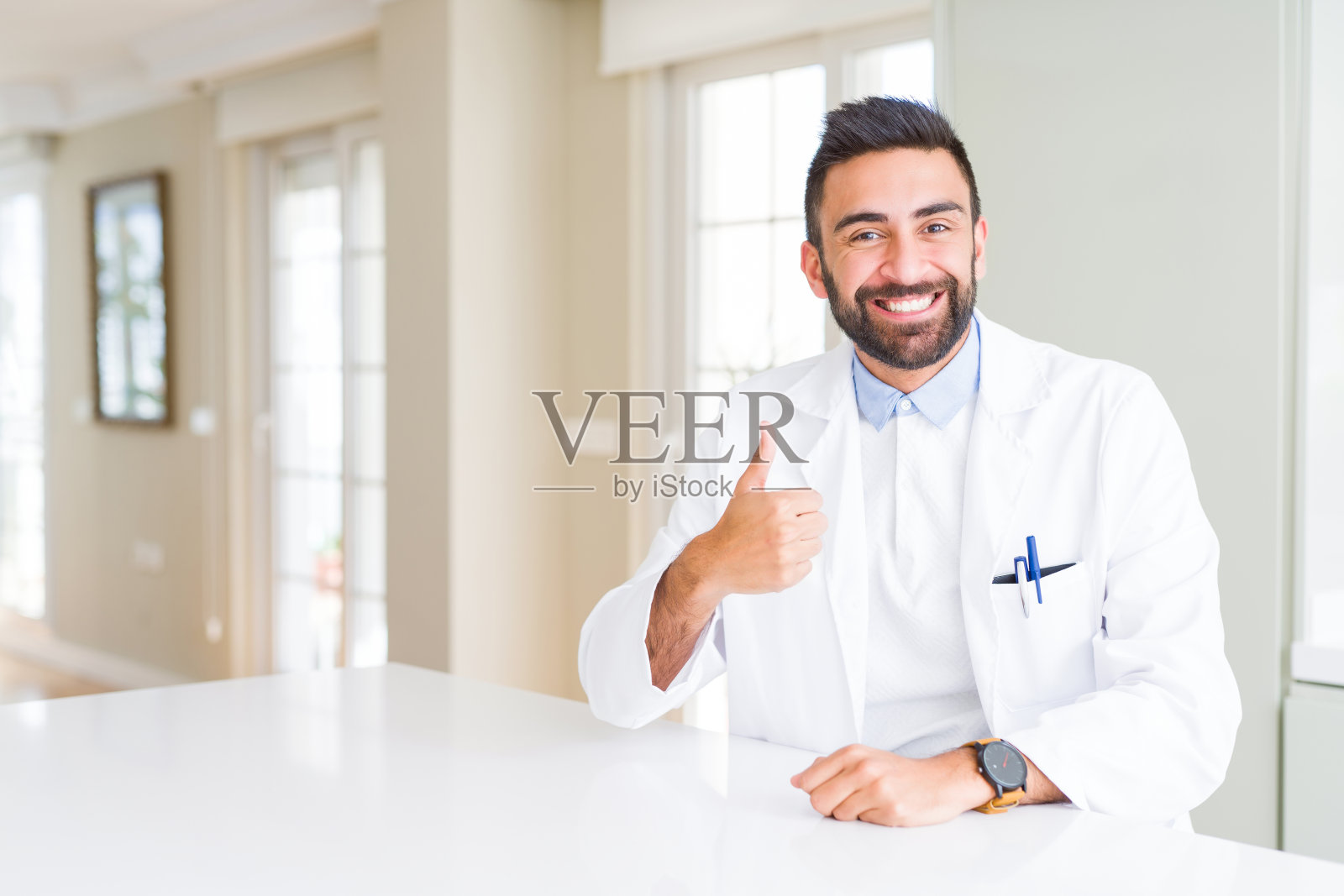 英俊的西班牙医生或治疗师穿着医疗外套在诊所做快乐的拇指手势与手。赞许的表情看着镜头，表示成功。照片摄影图片