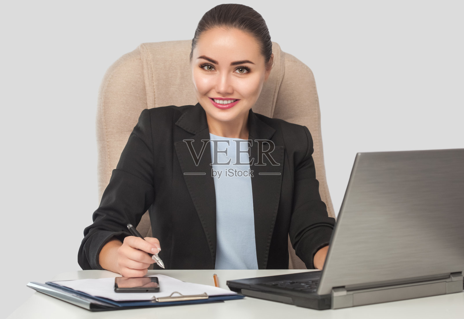 一个微笑的年轻女商人穿着正式的商务套装坐在办公室的桌子上用笔记本电脑工作。照片摄影图片