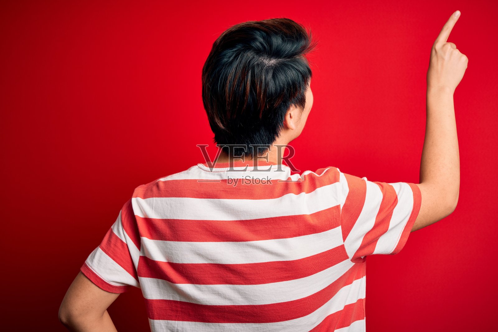 年轻帅气的中国男子穿着休闲条纹t恤站在红色背景下，用手指指着前方照片摄影图片