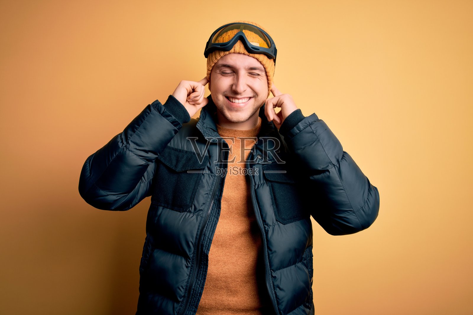 年轻英俊的白人男子戴着帽子，外套和滑雪眼镜冬天和下雪的天气用手指盖住耳朵，为大声的音乐噪音烦恼的表情。聋的概念。照片摄影图片