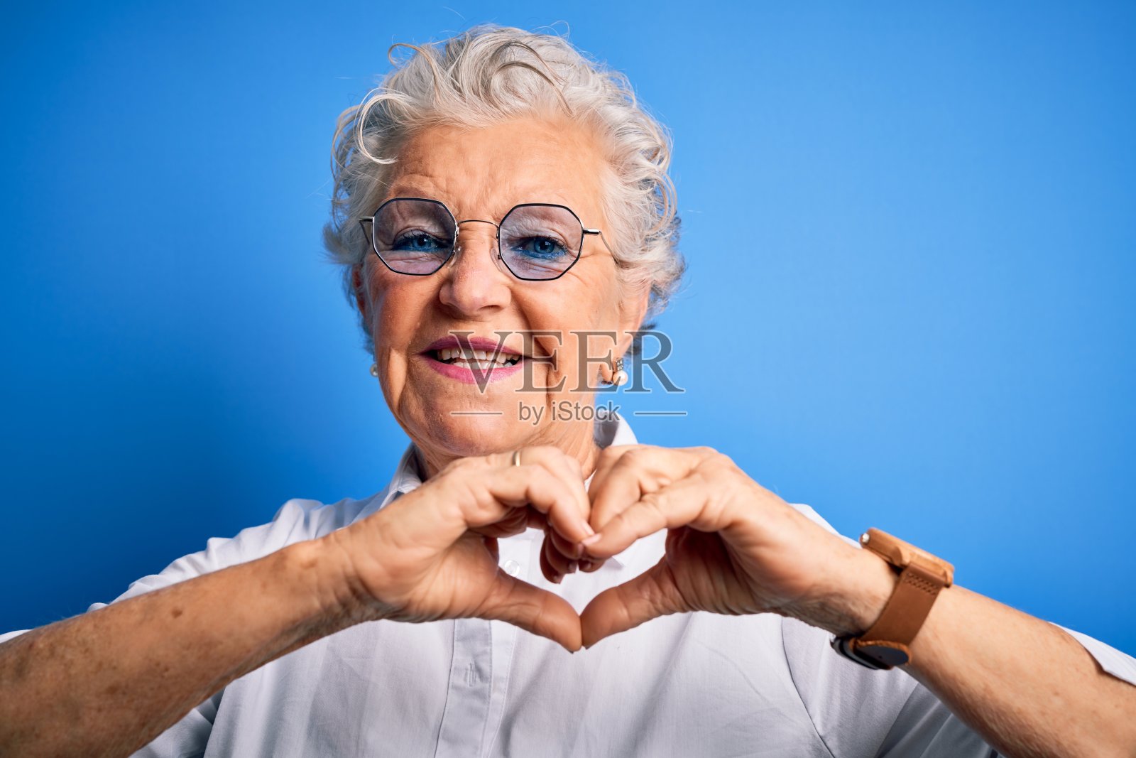 高级美丽的女人穿着优雅的衬衫和眼镜在孤立的蓝色背景微笑在爱显示心形符号和形状的手。浪漫的概念。照片摄影图片