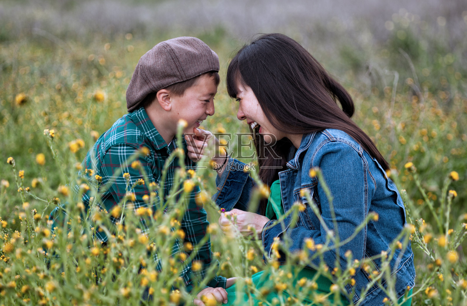 一位怀孕的亚洲母亲，一头乌黑的长发，穿着绿色的裙子和蓝色的牛仔夹克，她的儿子戴着棕色的报童帽，在一片长满黄花的绿色草地上大笑照片摄影图片