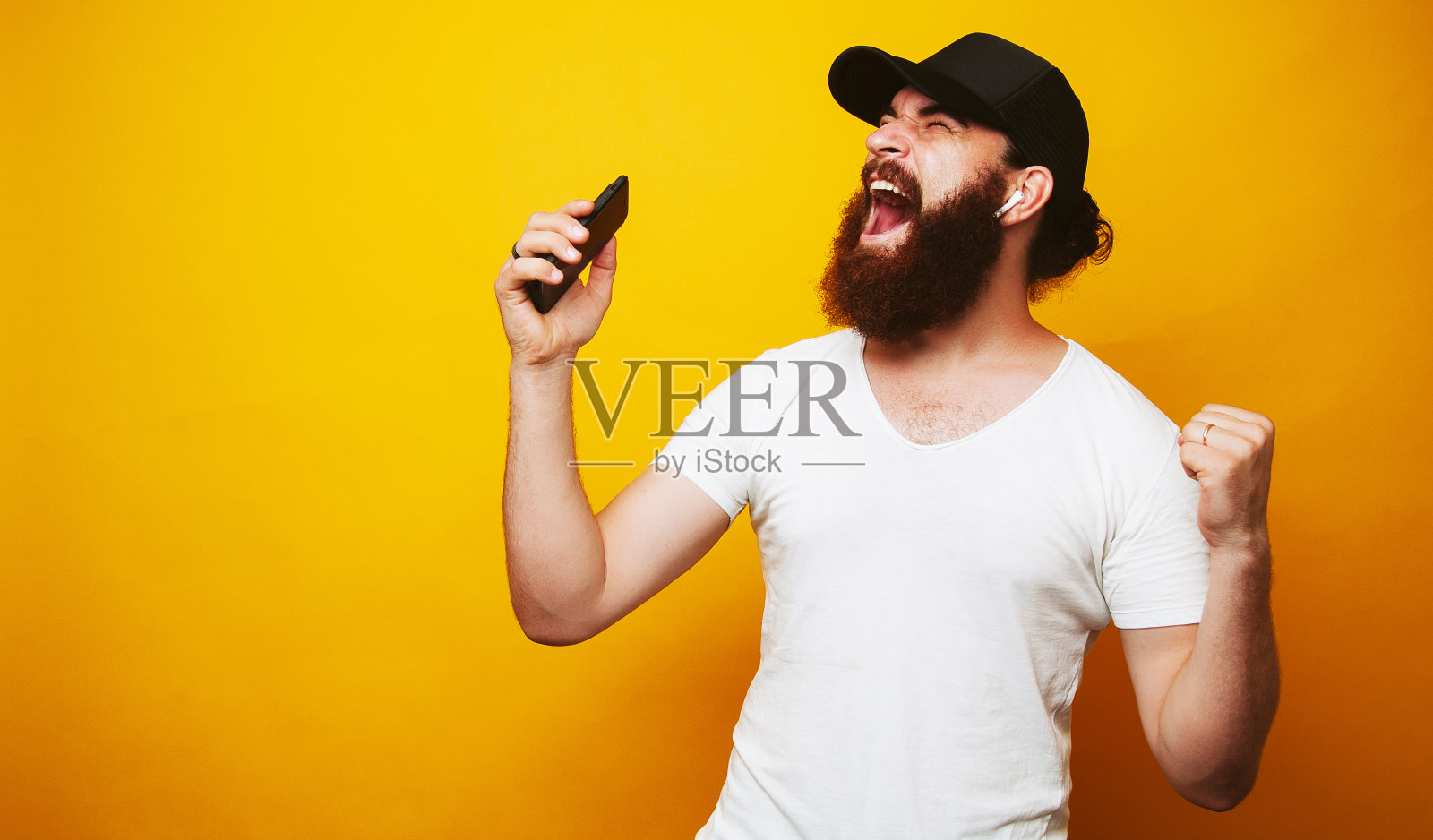 一个快乐的大胡子嬉皮士的肖像，一边唱歌一边听着音乐，用手机和无线耳机隔着黄色的墙照片摄影图片