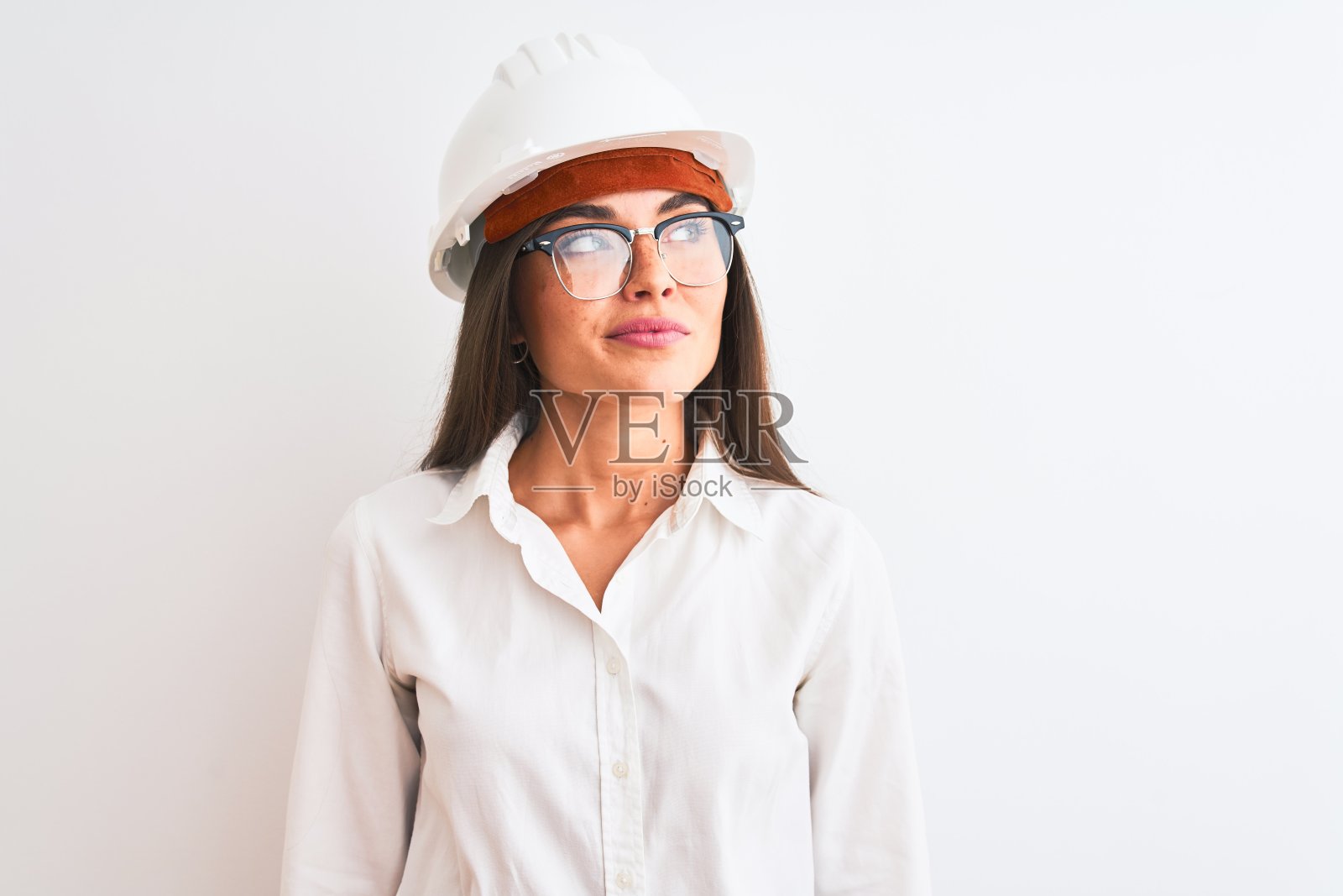 年轻美丽的建筑师女人戴着头盔和眼镜在孤立的白色背景微笑看向一边和凝视远离思考。照片摄影图片