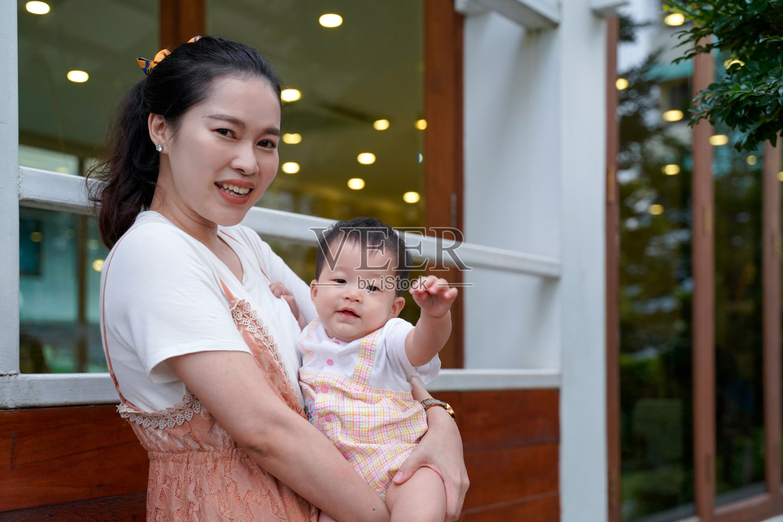 亚洲可爱的年轻母亲站在门口微笑着，抱着女儿在经典的玻璃木质入口背景下，宝宝在家门口或大楼外举手问候。照片摄影图片