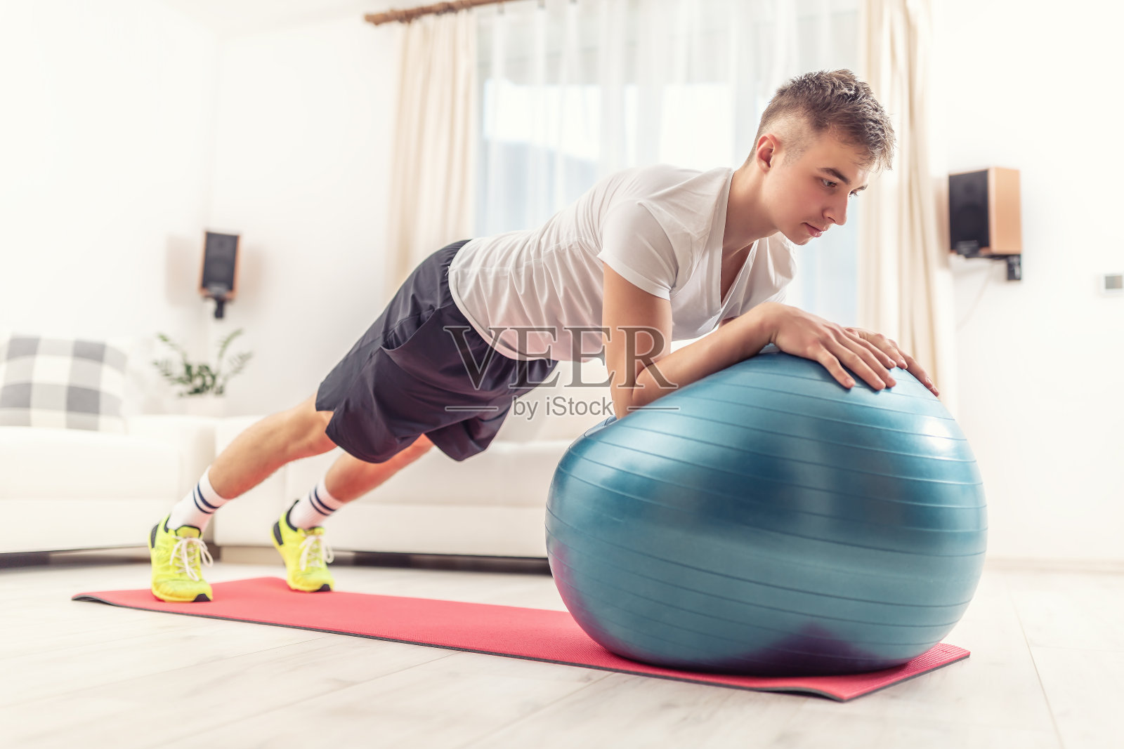 年轻人在家用一个蓝色的健身球和一个红色的垫子在客厅里做支撑姿势。照片摄影图片