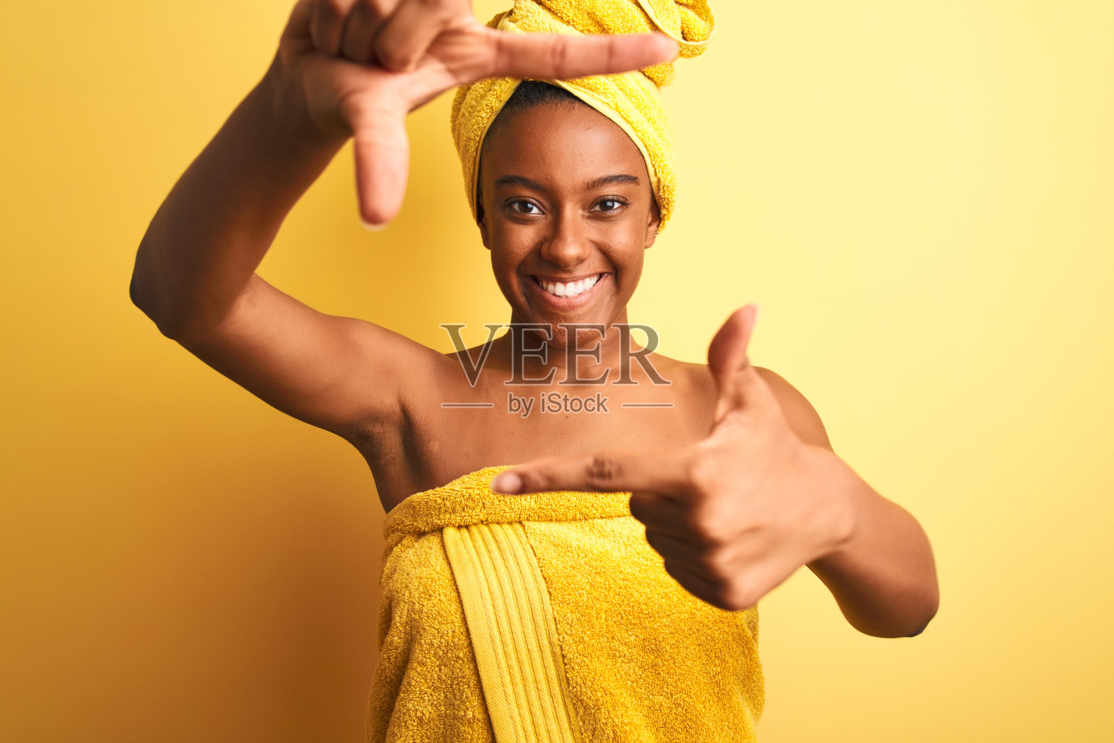 非洲裔美国妇女穿着毛巾淋浴后站在孤立的黄色背景微笑与手和手指框架与快乐的脸。创意和摄影概念。照片摄影图片