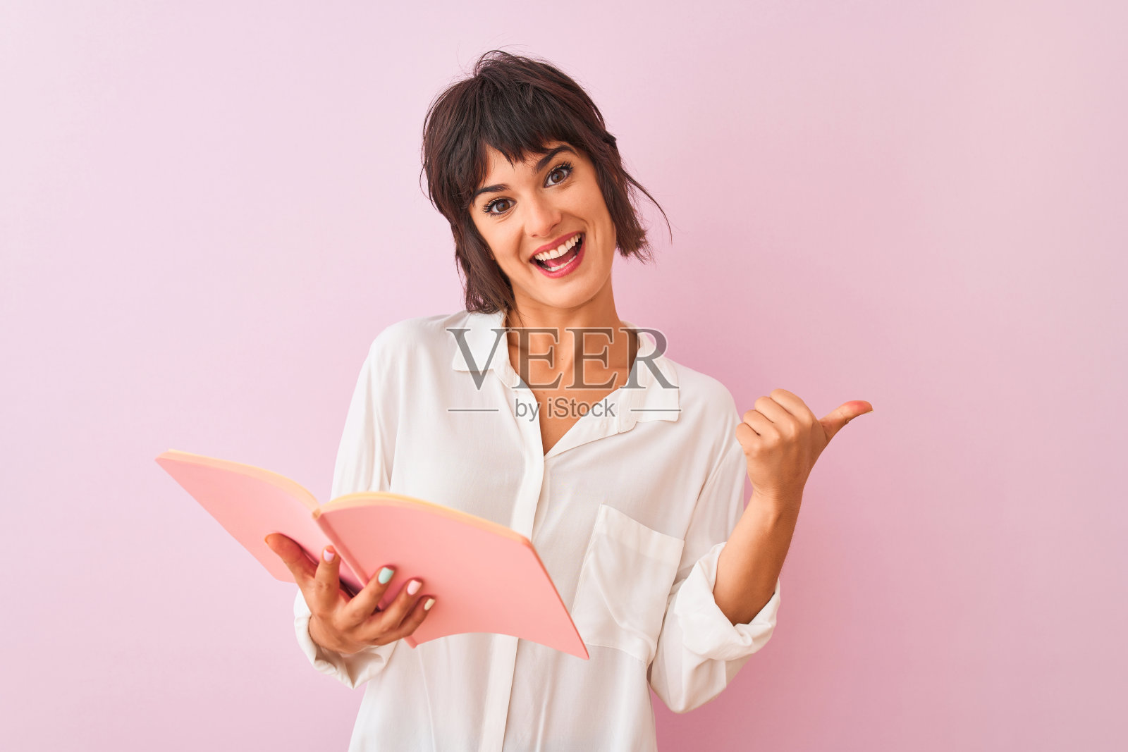 年轻美丽的女老师看书站在孤立的粉红色背景指向和显示与拇指向上的一边与幸福的脸微笑照片摄影图片