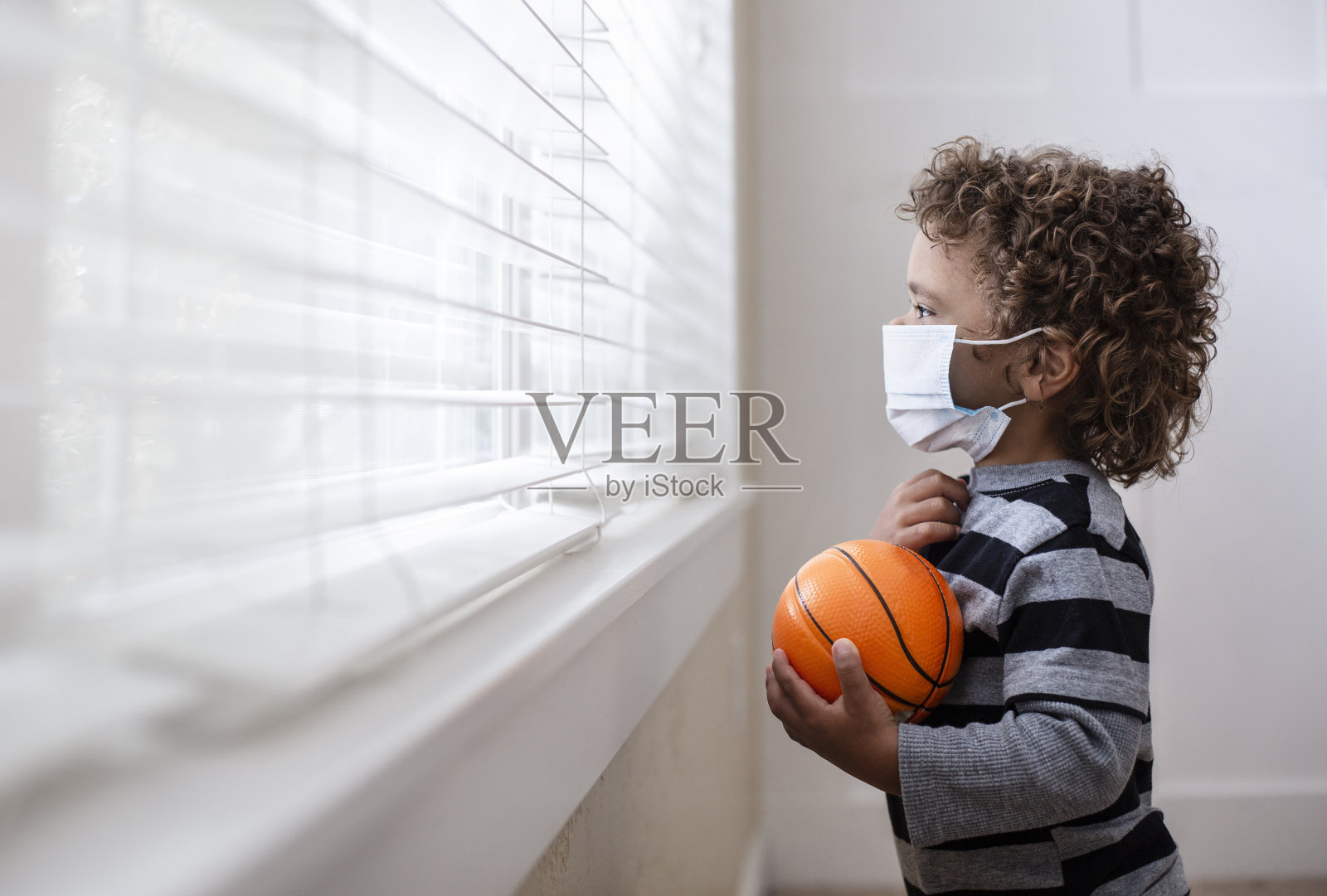 一名小男孩戴着防护面罩望着窗外，寻求防范COVID-19照片摄影图片