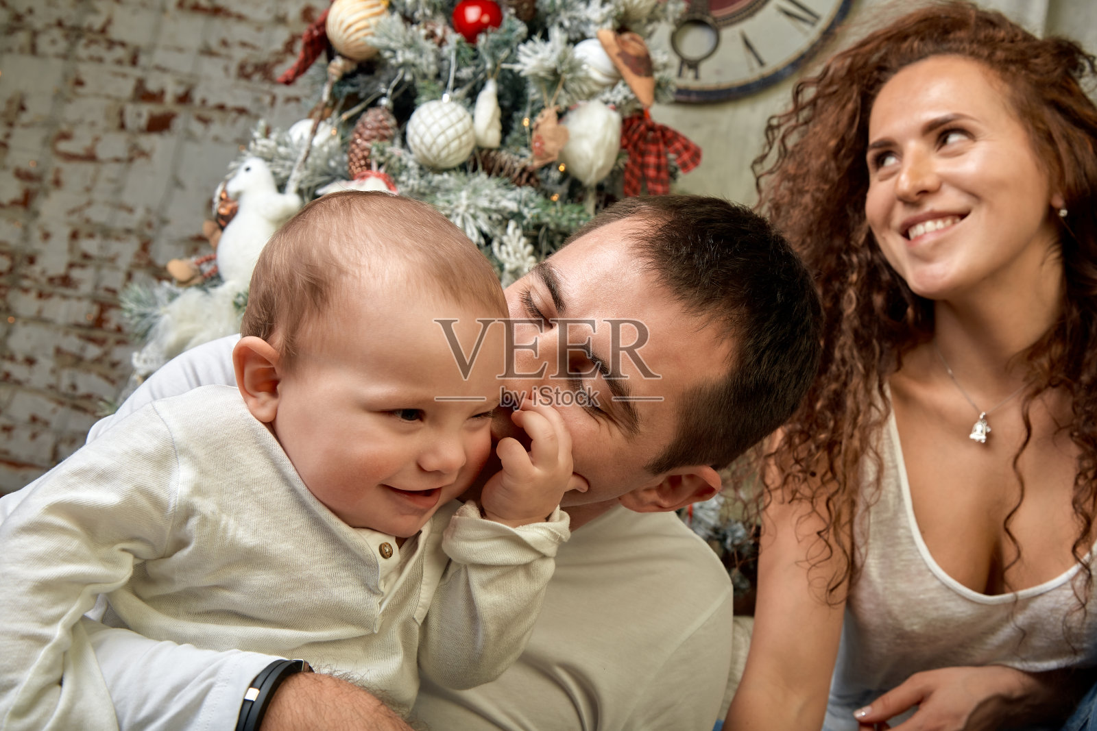 圣诞节的家庭。幸福。爸爸，妈妈的肖像。爸爸在家里靠近圣诞树和壁炉的地方亲吻宝宝，每个人都在微笑。新年快乐，合家欢乐照片摄影图片
