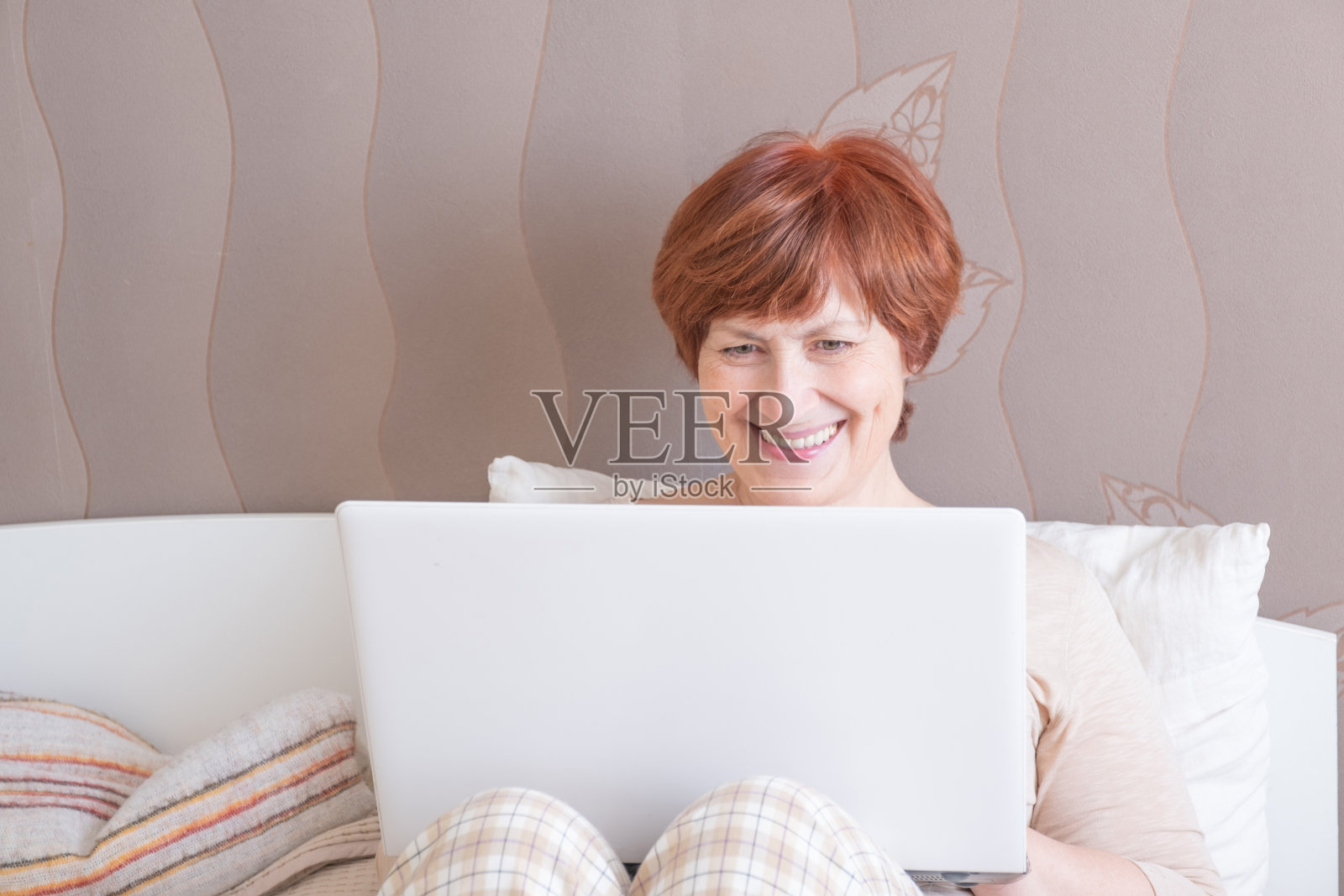 一位可爱的红短发高级女士正坐在床上拿着笔记本电脑，看着屏幕微笑着。照片摄影图片