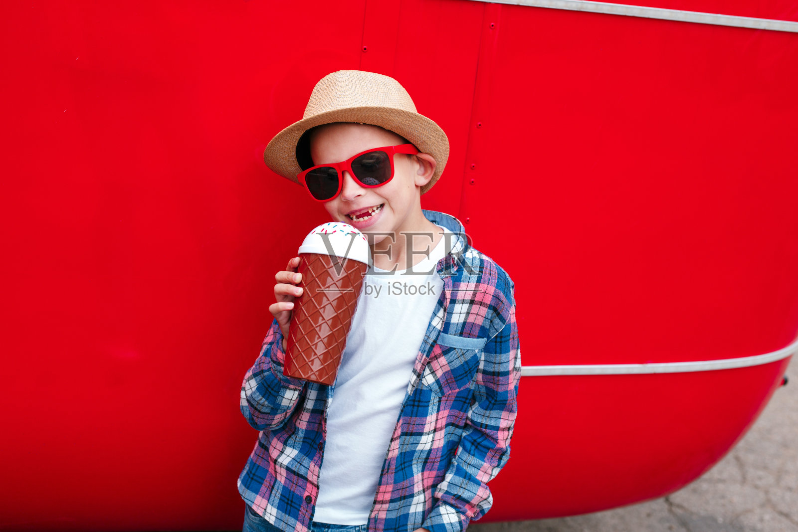 戴着墨镜和帽子的小男孩在红色的城市背景下喝着牛奶鸡尾酒照片摄影图片