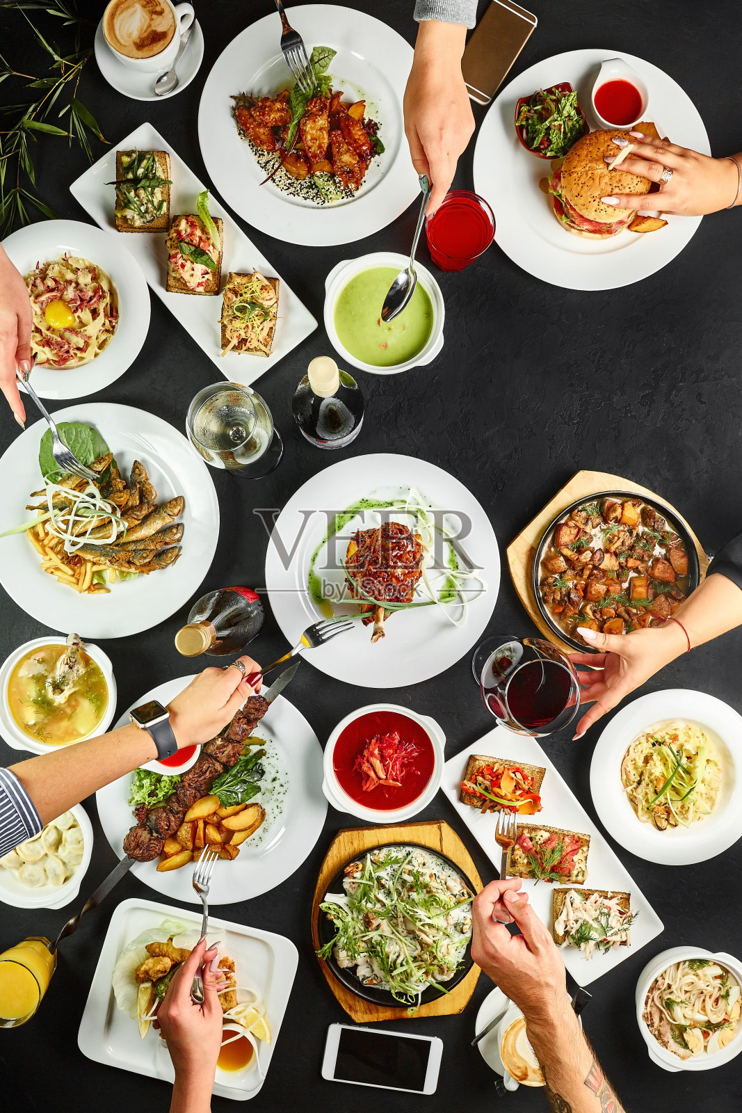各种食物组成的菜肴和手与刀叉在黑色的石板背景。与亲朋好友共进晚餐。餐厅吃饭的概念照片摄影图片