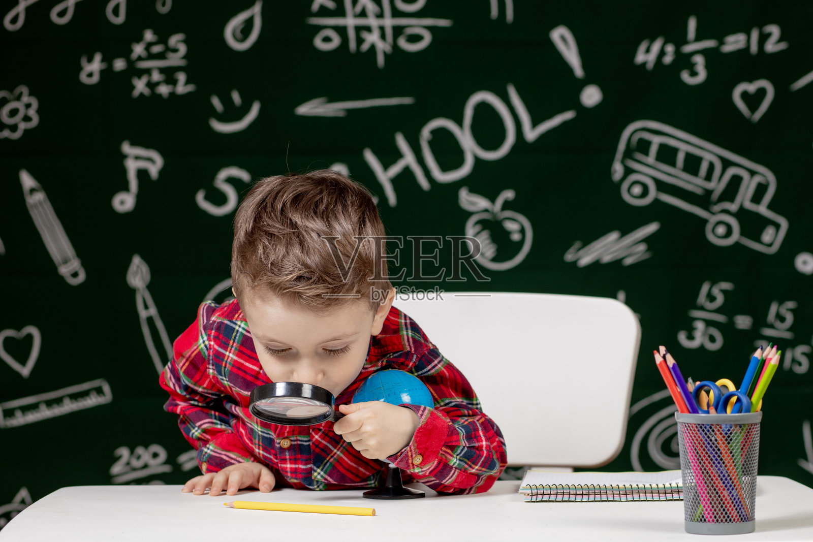可爱聪明的男孩坐在书桌前，手里拿着放大镜。孩子正在读一本以黑板为背景的书。准备好学校。回到学校照片摄影图片