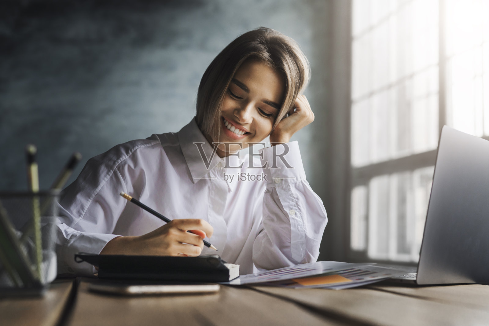 微笑的女孩坐在书桌前，用钢笔在抄写本上做笔记。女学生在笔记本电脑前学习照片摄影图片