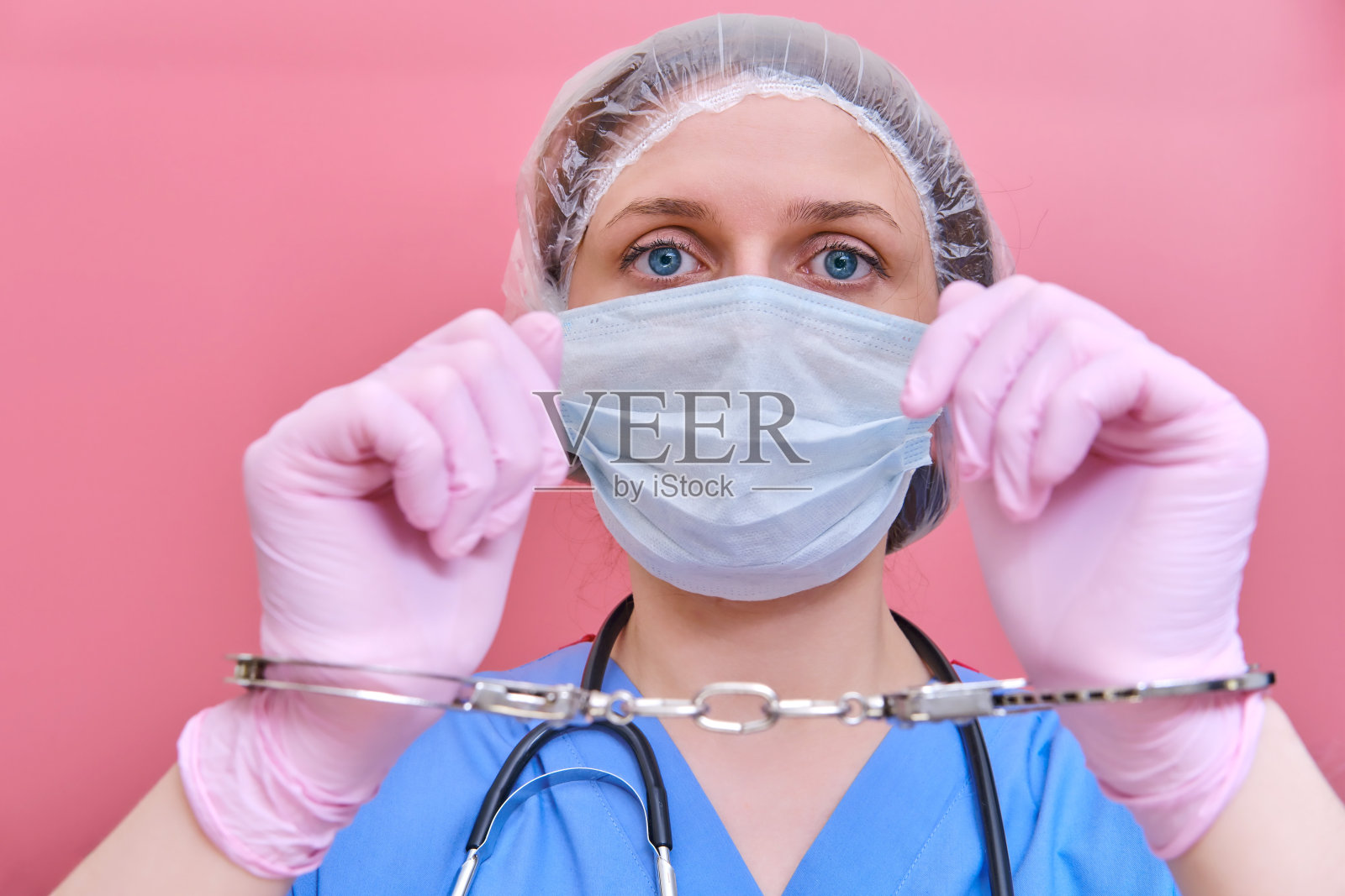 一个带着手铐的医用面具的女医生的肖像，特写。护士身穿蓝色制服，戴着防护手套，背景为粉红色。照片摄影图片